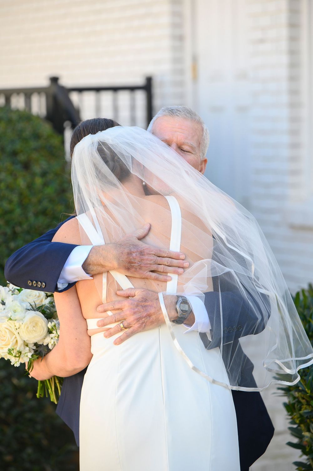 A tender embrace at an outdoor wedding ceremony with the bride wearing a flowing white gown and veil holding a bouquet.