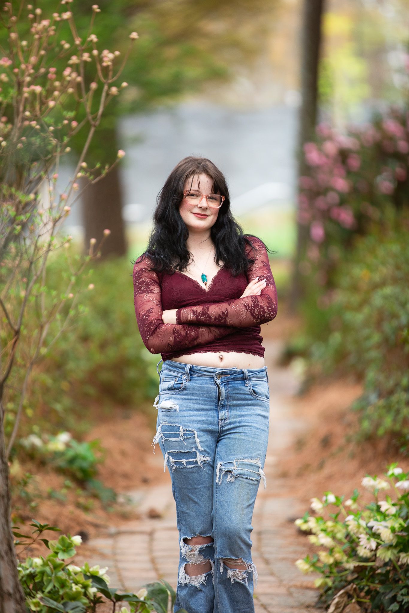 Woman in burgundy lace top and jeans stands with arms raised overhead in a lush green outdoor park setting.