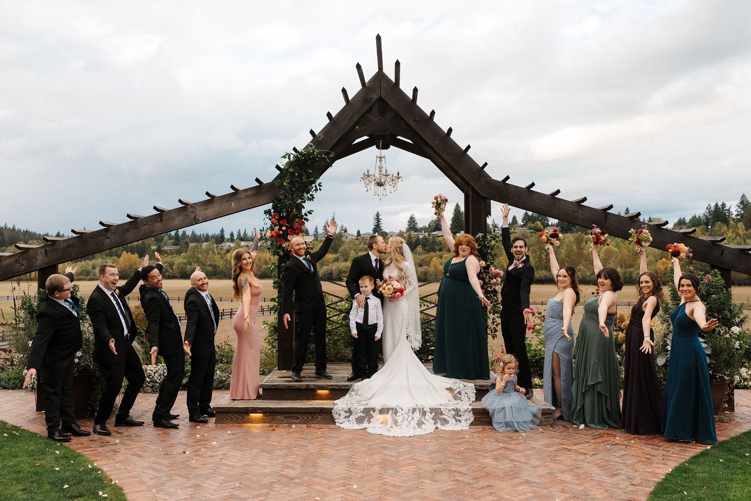 Wedding party members line up for a panoramic group photo on a grassy hill with mountains in the background.