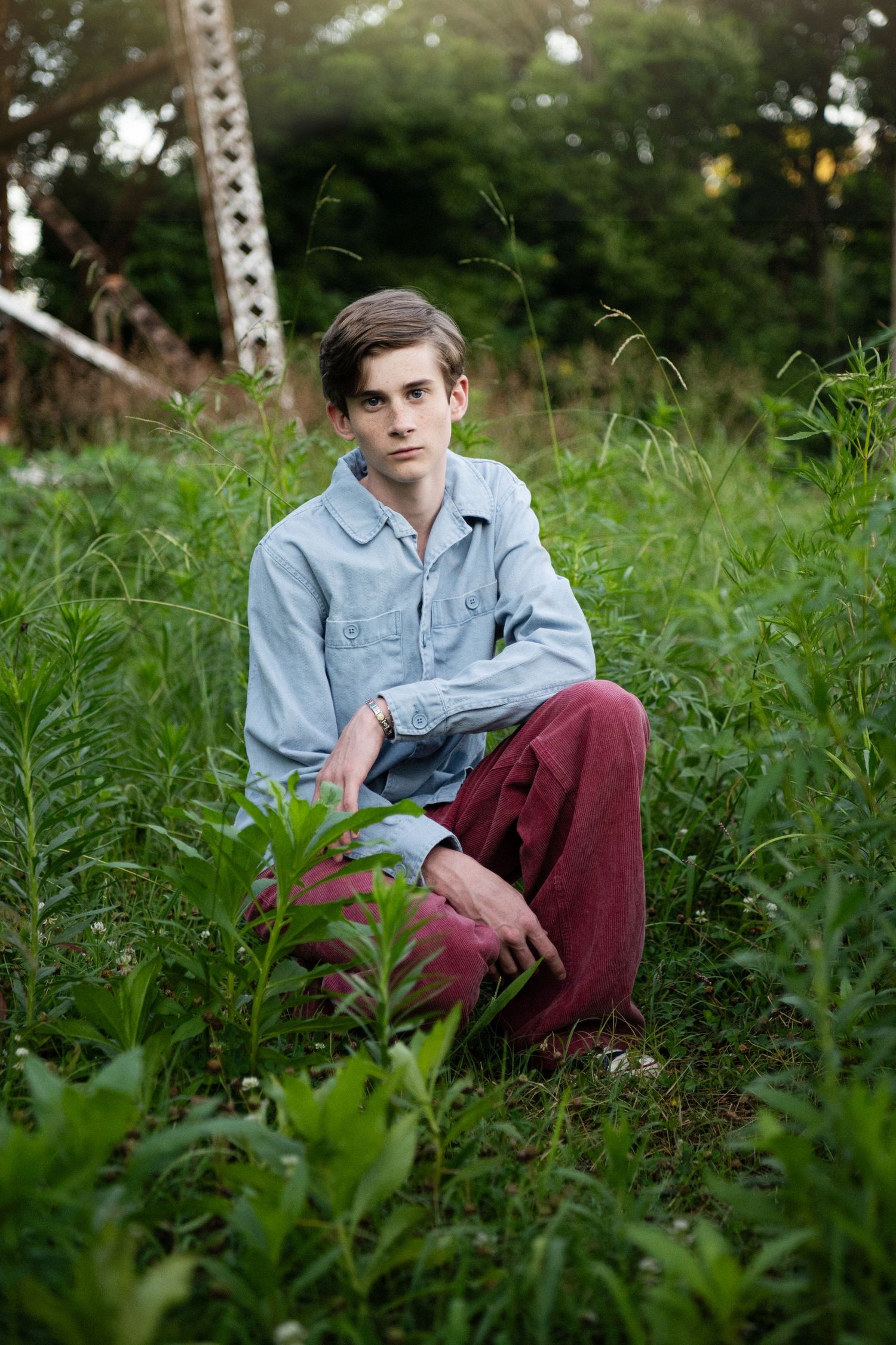 A person in a denim jacket and burgundy pants sits among tall green grass in an outdoor setting.