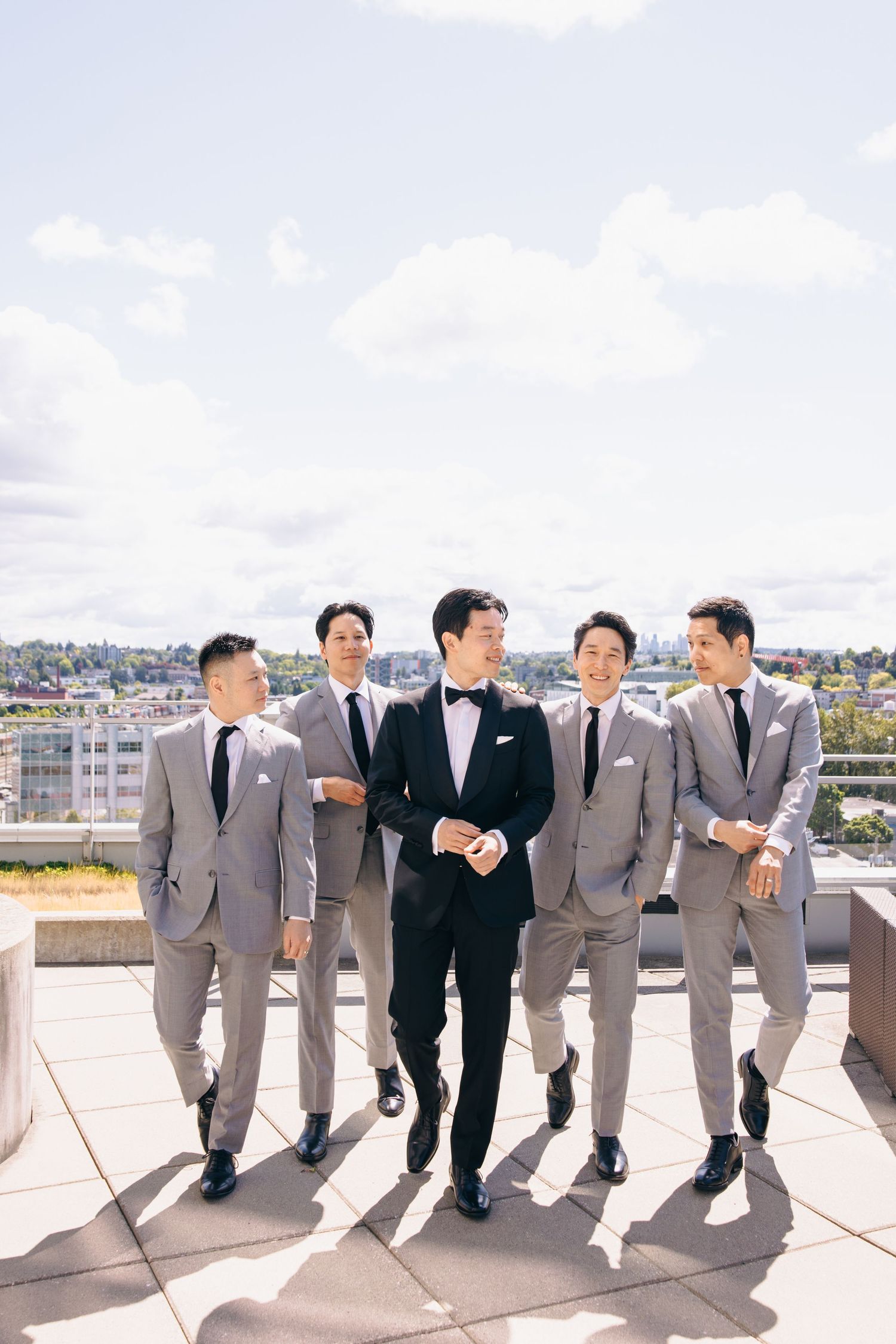 A group of groomsmen in gray suits and black tuxedos lean casually against a rooftop wall on their wedding day.