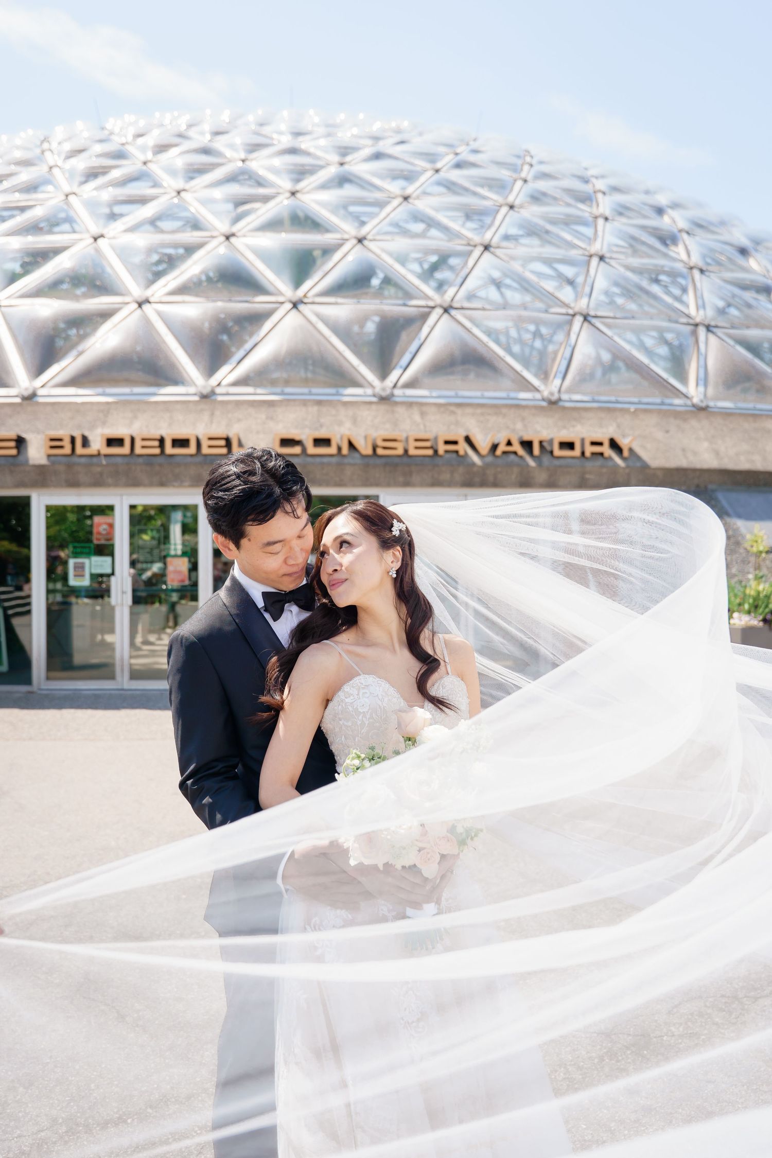 Romantic wedding couple embraces under a modern glass conservatory dome with flowing white veil in the breeze.Queen Elizabeth Park Wedding