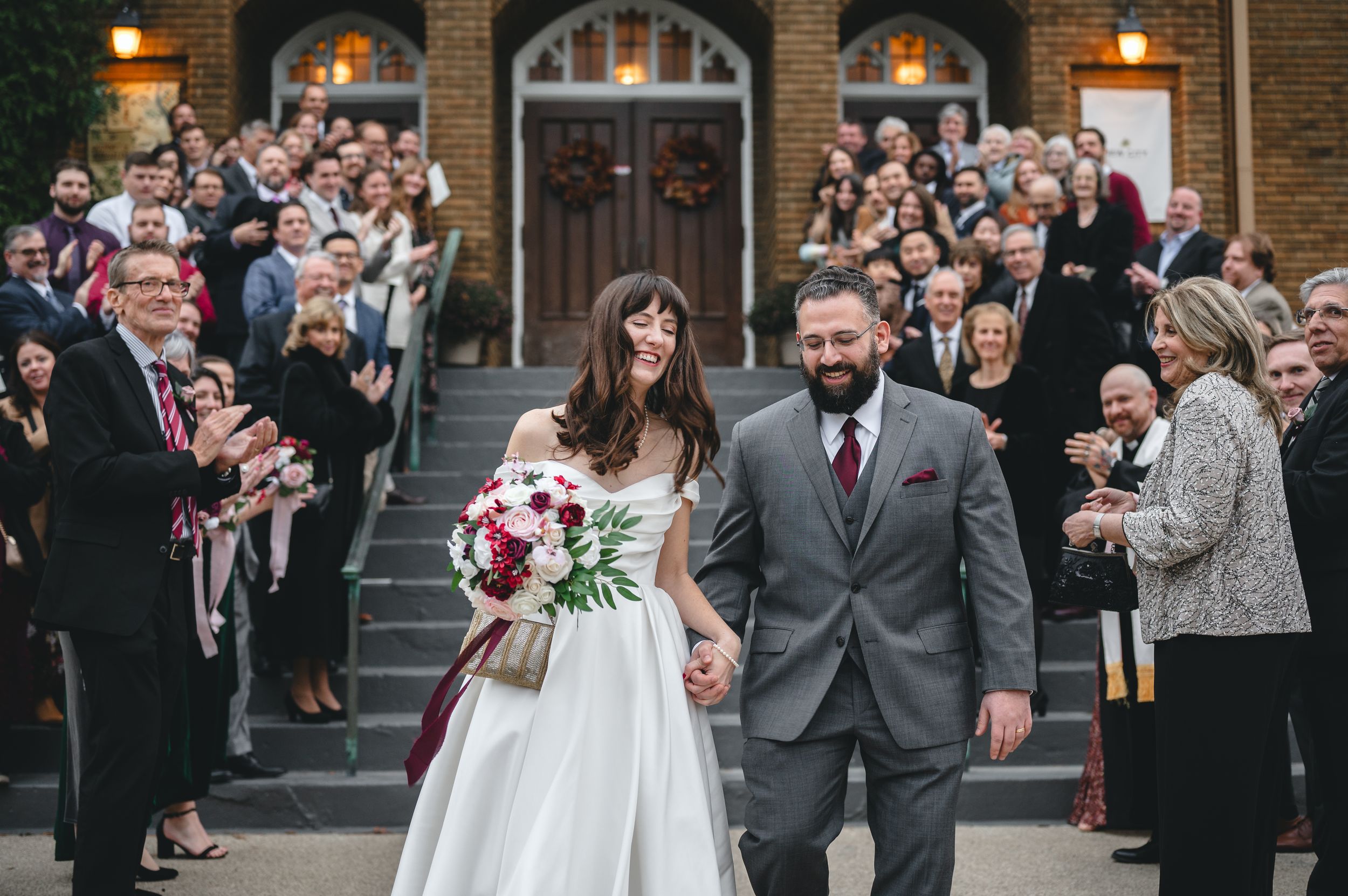 Wedding guests celebrate and cheer as a couple exits their ceremony at a historic brick church venue.