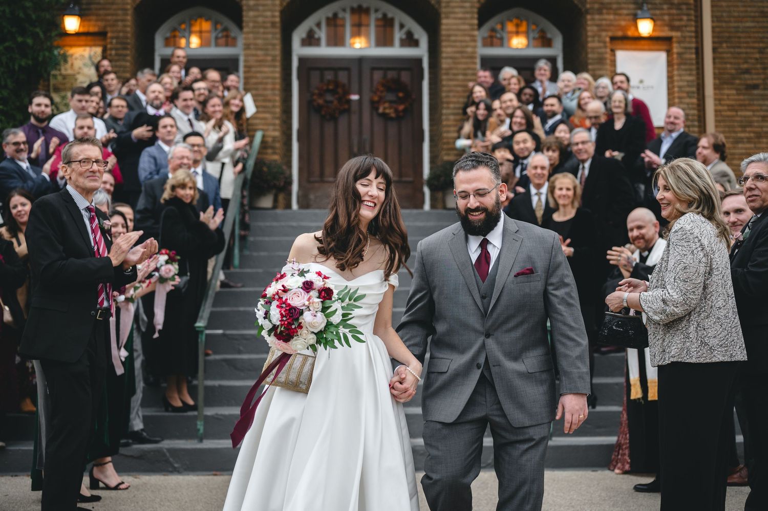 Wedding guests celebrate and cheer as a couple exits their ceremony at a historic brick church venue.