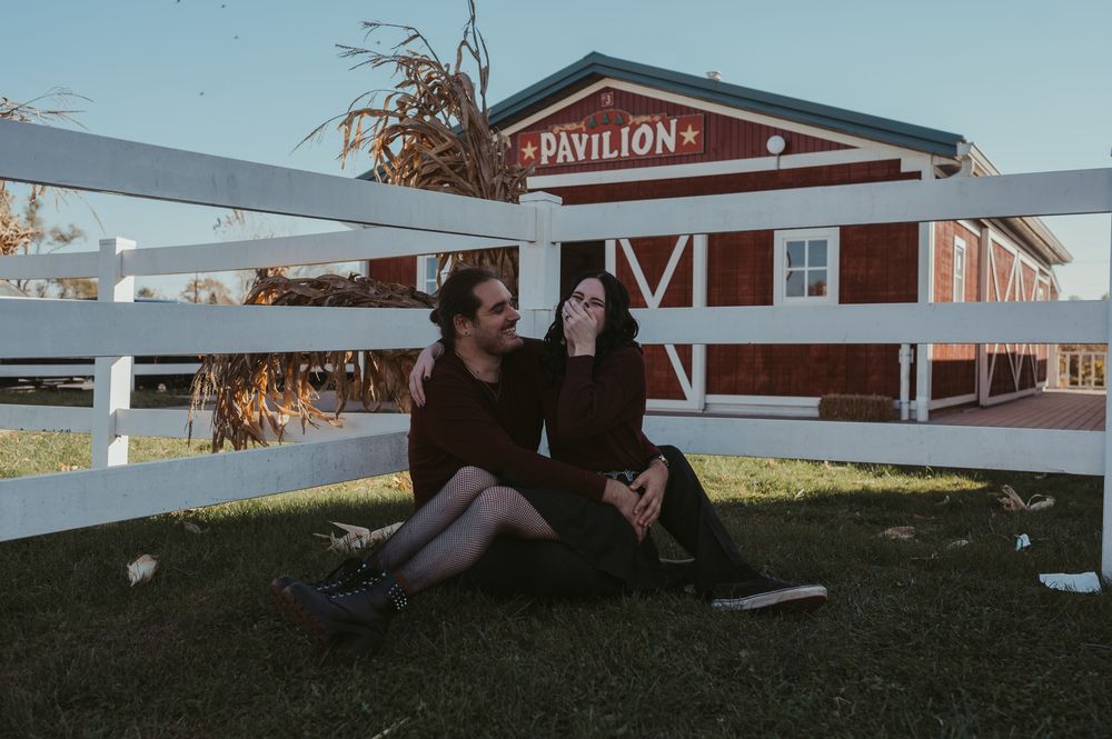 Couple sitting on grass in front of a red Pavilion barn with white fence during autumn.