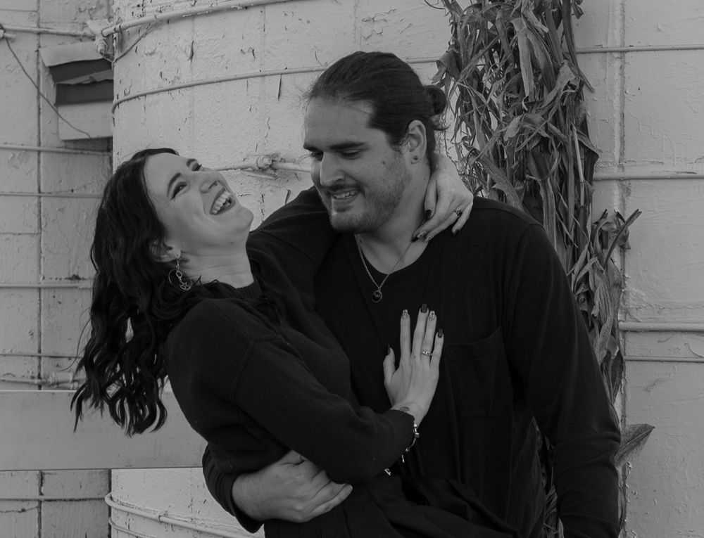 Black and white photo of a laughing couple embracing outdoors against a rustic wall.