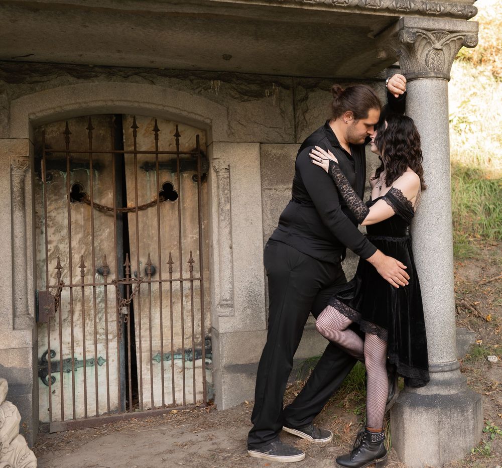 Couple in black gothic attire embracing by a stone mausoleum with iron gate.