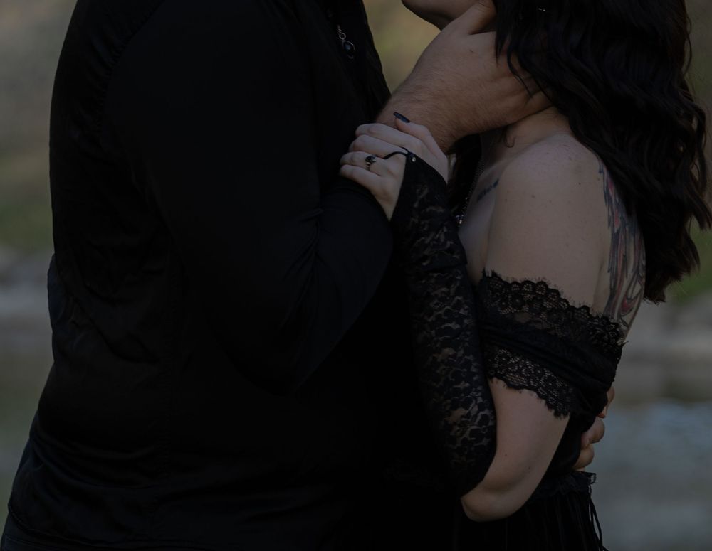 Couple in black attire sharing a romantic embrace, woman wearing lace off-shoulder dress outdoors.