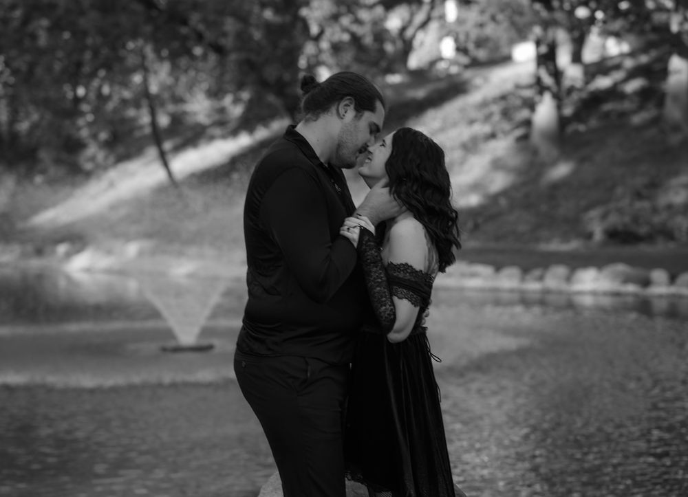 Black and white photo of a couple sharing a romantic kiss near a fountain in a park.