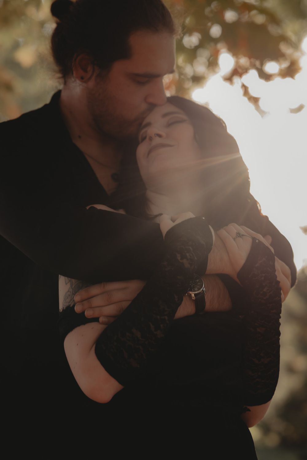 Couple embracing in golden backlight, man kissing woman's forehead, both wearing black in an outdoor autumn setting.