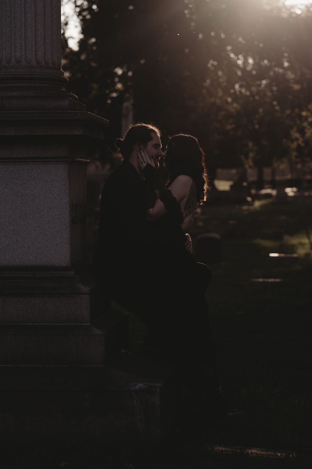 Two people sharing a romantic kiss beside a stone monument in a sunlit park at golden hour.