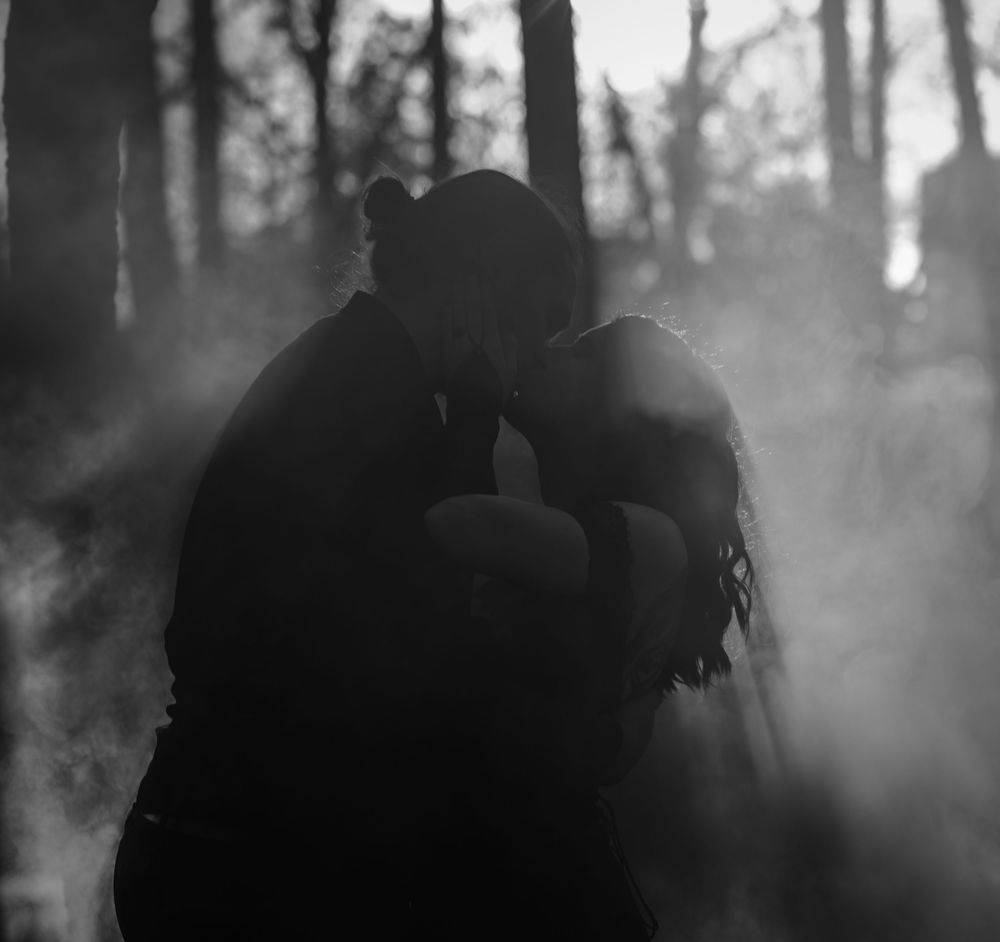 Black and white photo of a couple kissing in a misty forest, surrounded by atmospheric smoke and bare trees.