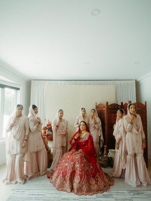 Wedding group photo with bridesmaids in light beige and bride in vibrant red traditional attire.