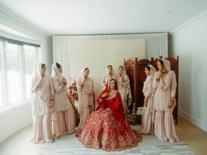 A bride in a vibrant red lehenga sits surrounded by bridesmaids wearing soft pink traditional outfits in a bright room.