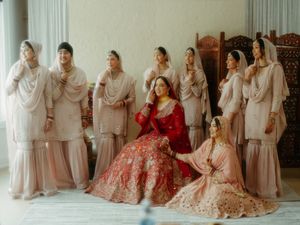 Wedding party lined up against white backdrop with bride in red lehenga and attendants in neutral tones.