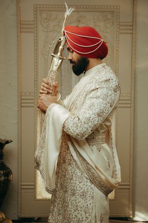 A person wearing a white embroidered traditional Sikh wedding outfit and red turban poses in an elegant room.