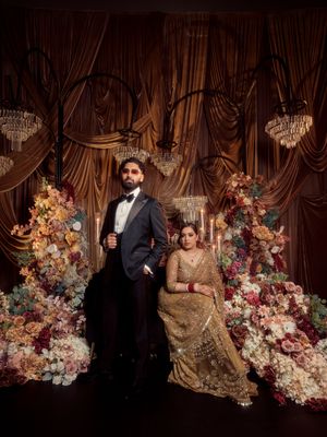 Elegant couple poses against a dramatic backdrop of cascading flowers and draped curtains.