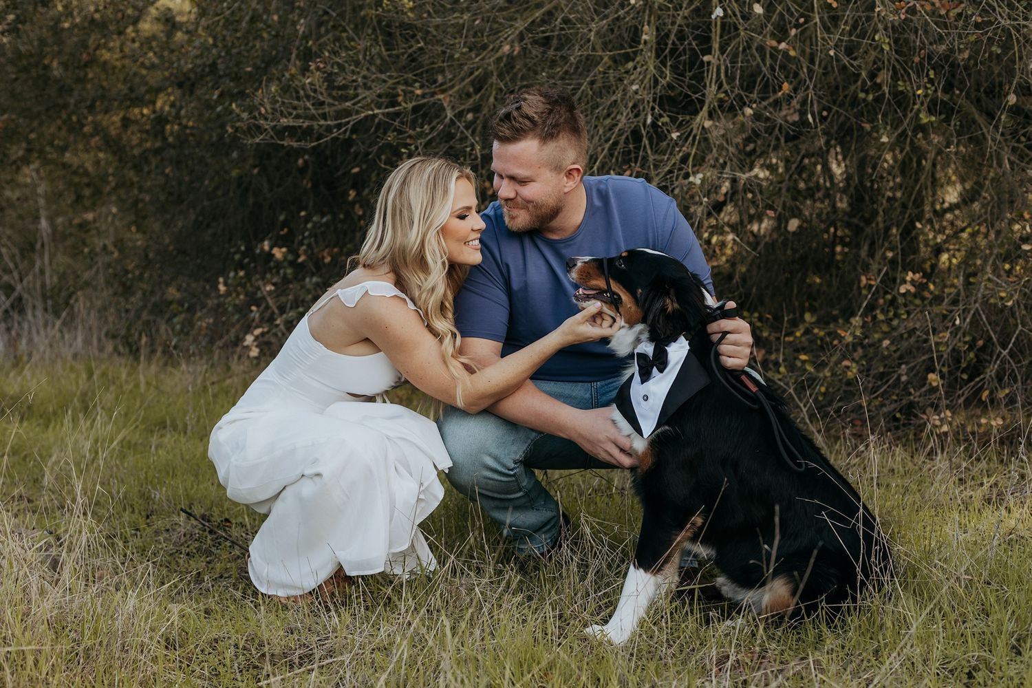 A couple sits together on grassy ground with their black dog during an outdoor photo session in San Diego.