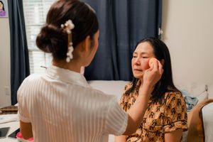 Mother of Bride in floral dress having makeup applied by Vietnamese bride during Dallas wedding preparation indoors.