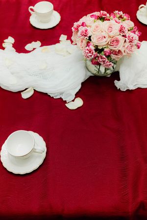 Red tablecloth with white teacups, saucers, rose petals, and pink floral centerpiece for Vietnamese tea ceremony.