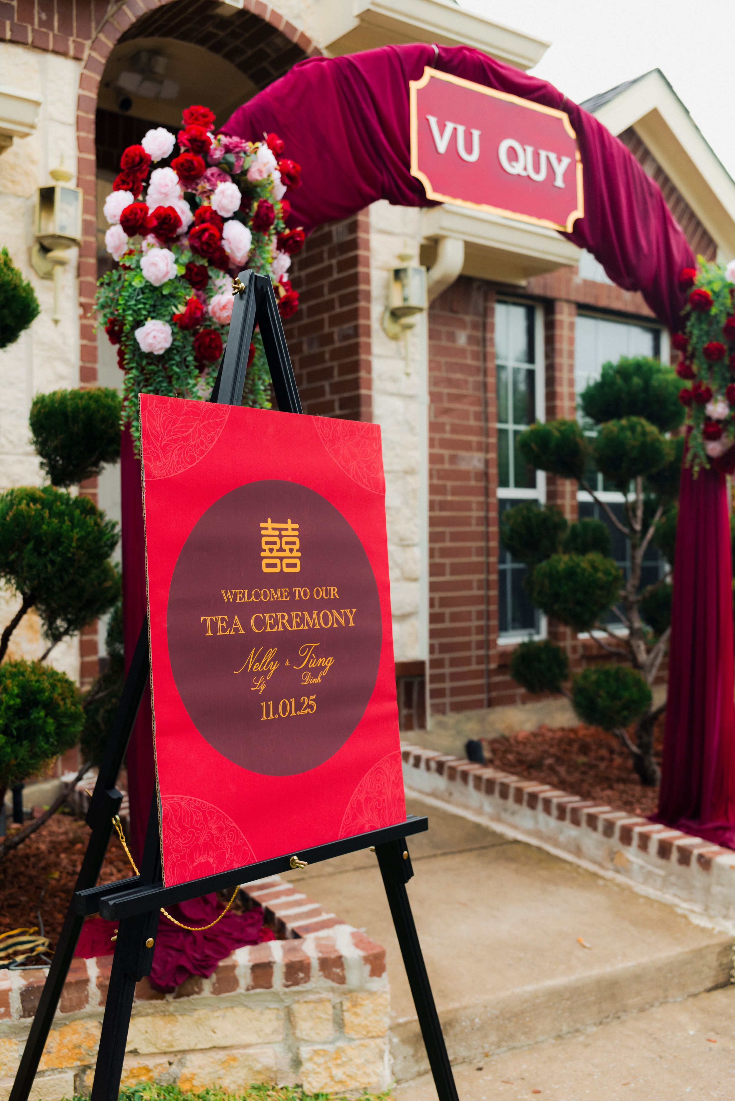 Red welcome sign for Vietnamese tea ceremony Vu Quy on easel with floral arch and brick house backdrop.