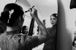 Bride applying makeup while looking in mirror during black and white wedding preparation photo.