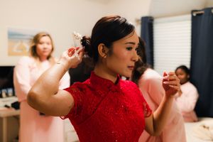 Bride in red lace qipao cheongsam dress getting hair accessory placed by bridesmaids in pink robes.