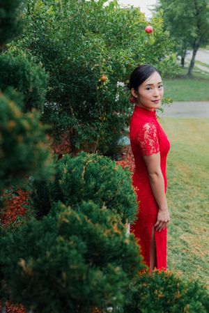 Elegant Vietnamese bride in red lace qipao cheongsam posing outdoors among lush green garden shrubs.