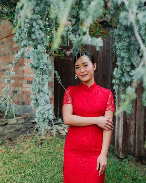 Beautiful Vietnamese bride in red lace cheongsam dress posing under weeping evergreen branches outdoors.