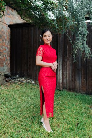 Vietnamese bride in red lace qipao cheongsam dress posing in garden near wooden fence and brick wall.