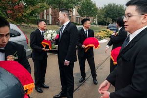 Groomsmen in black suits carry red traditional Vietnamese wedding gifts outside.