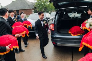 Groom in tuxedo greets groomsmen carrying red traditional Vietnamese wedding trays by car.