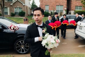 Groom in tuxedo holds white bouquet with groomsmen carrying red trays behind him.