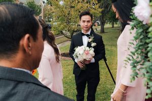 Groom in black tuxedo holds bouquet while greeting bridesmaids in pink ao dai outdoors.