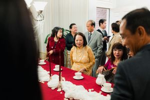 Guests gather around red-draped table with tea set at Vietnamese wedding reception.