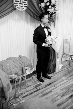 Groom in tuxedo holds bouquet beside double happiness symbol at wedding altar.