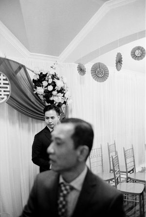 Groom stands near floral arrangement beneath double happiness symbol at wedding venue.