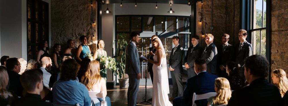 A couple shares their first dance in a rustic industrial venue with large windows and guests watching.