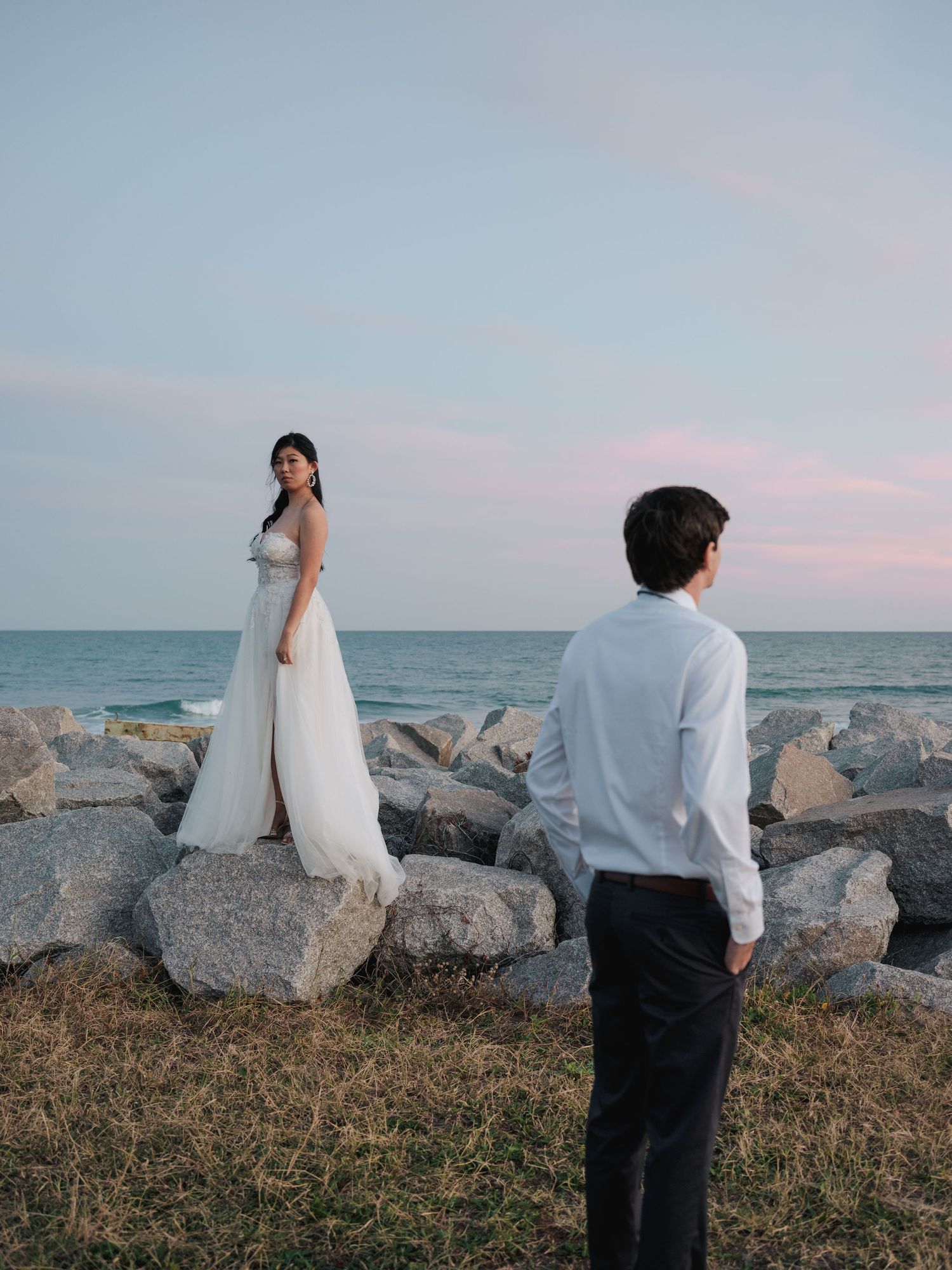 A couple in wedding attire stands on rocky shoreline against pastel sunset sky and ocean backdrop.