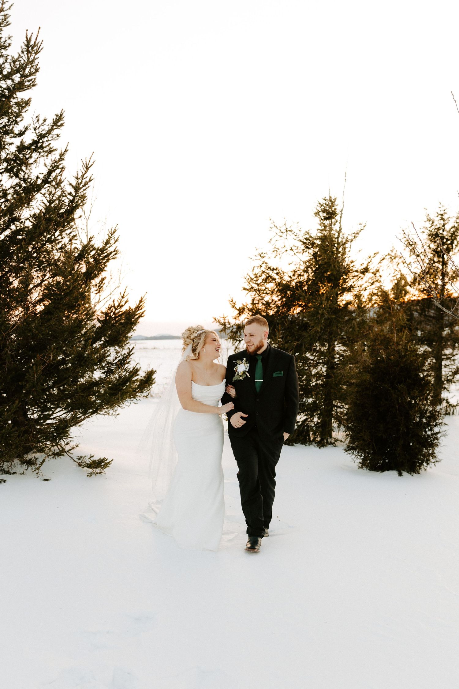 Bride and groom walk hand in hand through snowy pine tree landscape during winter wedding portraits.
