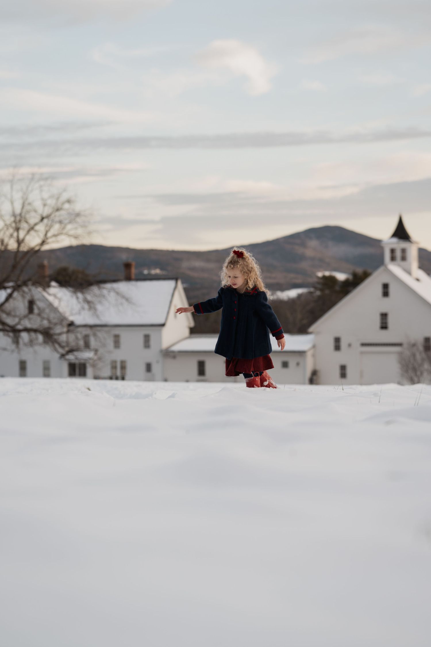 A young girl in a black coat and red dress walks through snow near white farm buildings and mountains.