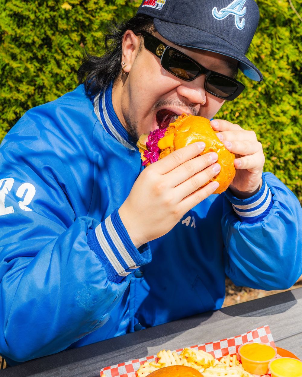 Person in blue sweatshirt and baseball cap takes multiple bites of a large sandwich at an outdoor table.