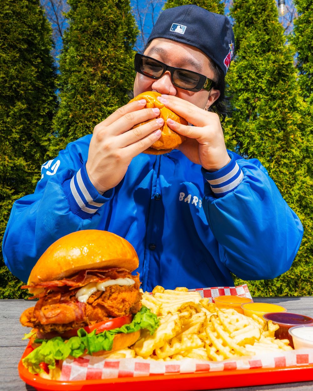 Someone in a blue jacket takes a big bite of a massive burger with crinkle cut fries on a red tray.