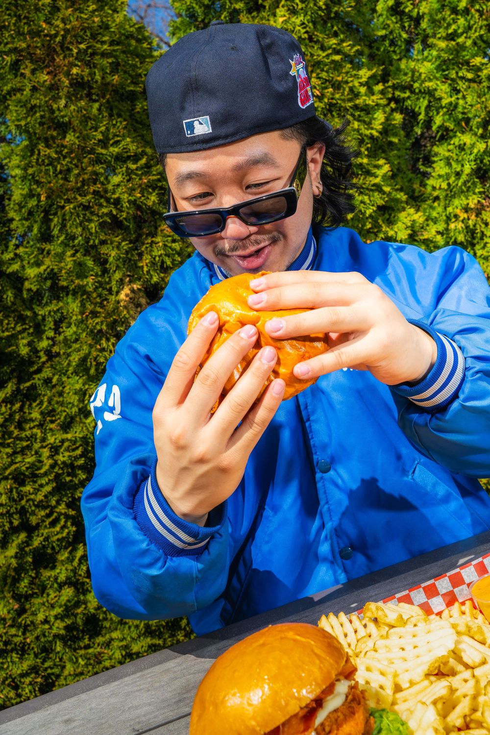 Someone in a blue jacket and baseball cap enjoying a large burger and fries outdoors on a sunny day.