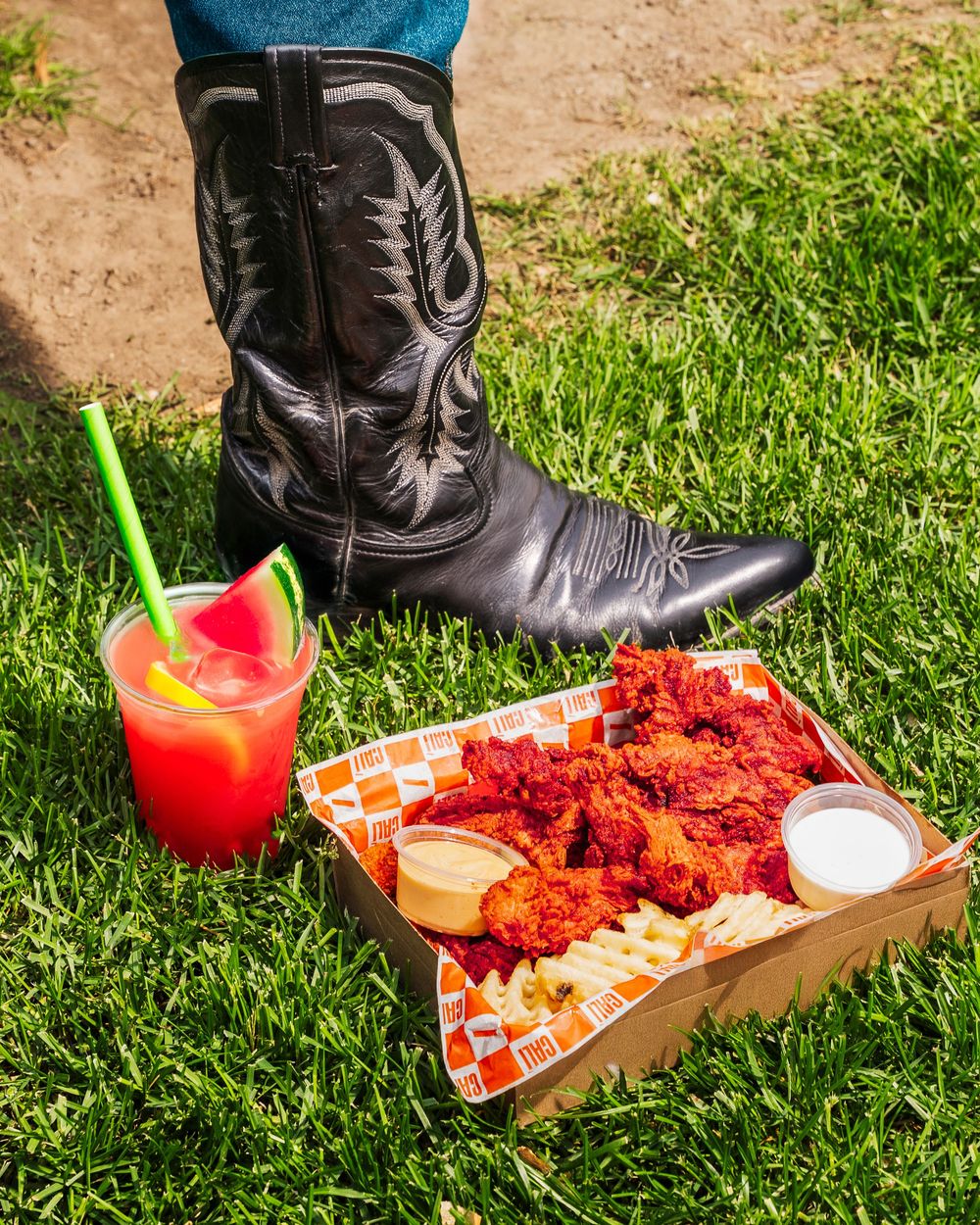 Black leather cowboy boots next to spicy buffalo wings and a tropical cocktail on grass.