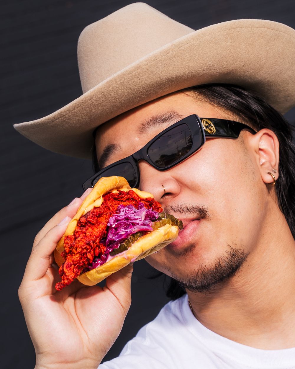 A person wearing a cowboy hat takes a bite of a colorful hot dog with vibrant toppings against a black background.