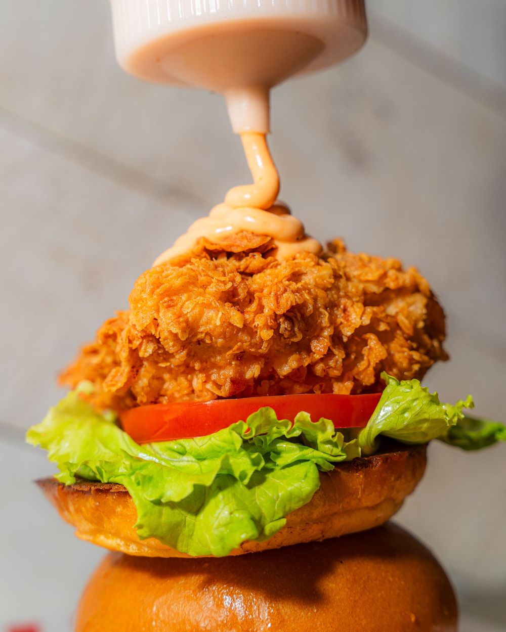 Crispy fried chicken sandwich with lettuce and tomato being assembled on a brioche bun against a light background.
