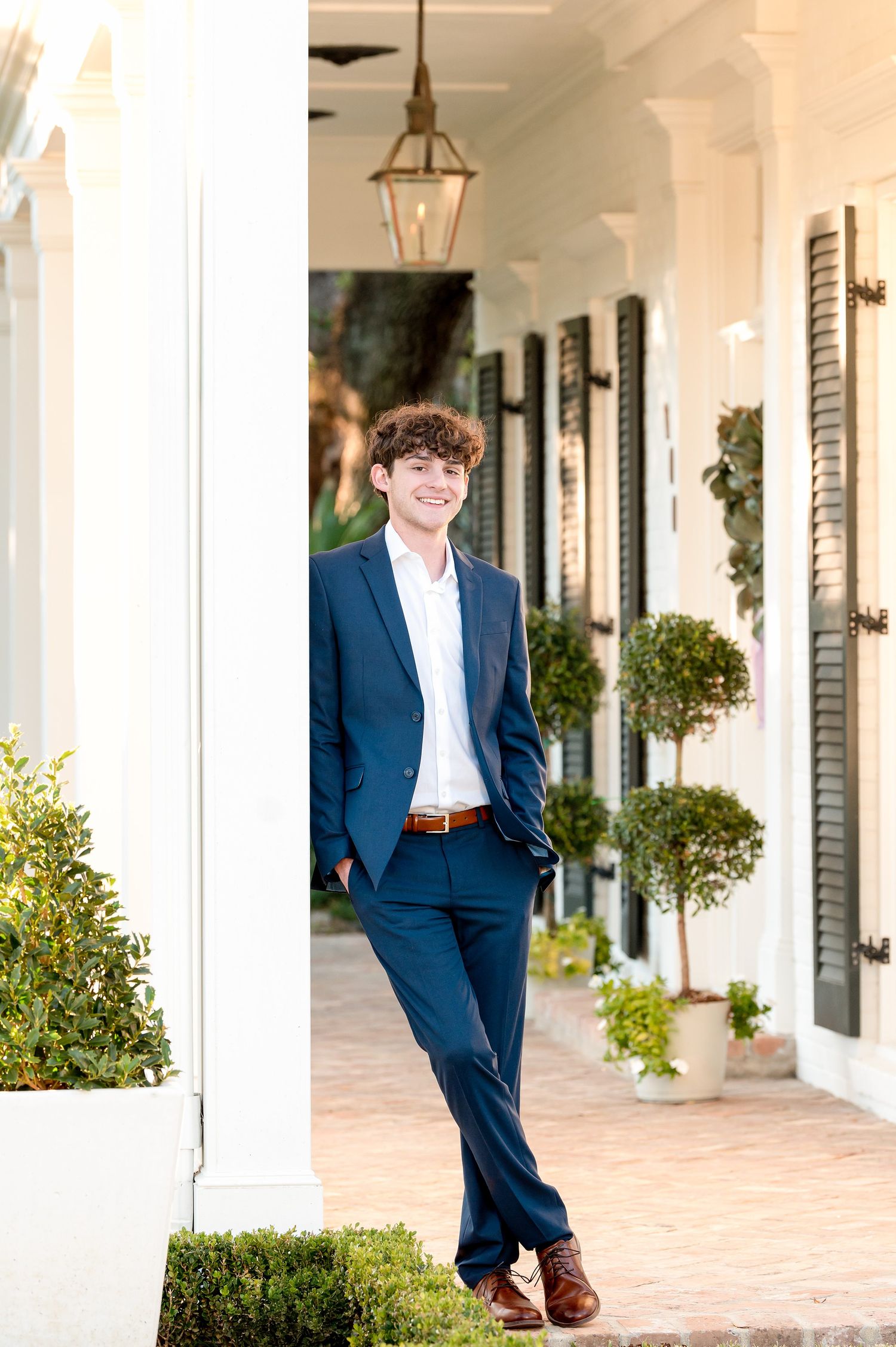 A person in a navy suit stands in a covered outdoor walkway with white columns and potted plants.