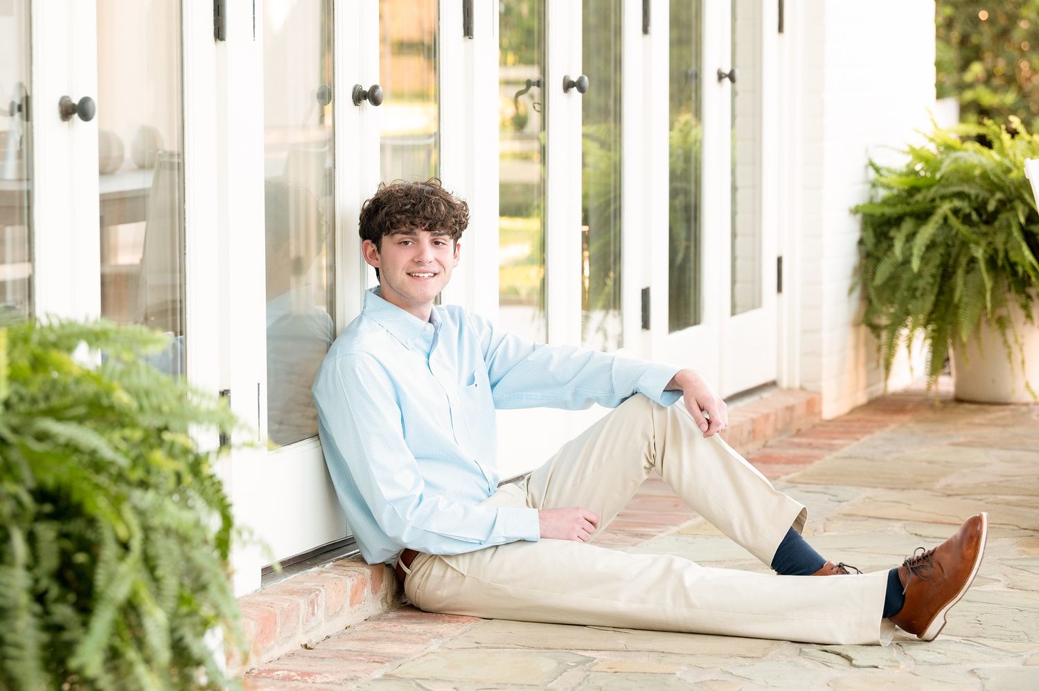 A person in a blue shirt sits casually on a white porch railing with plants and greenery in the background.
