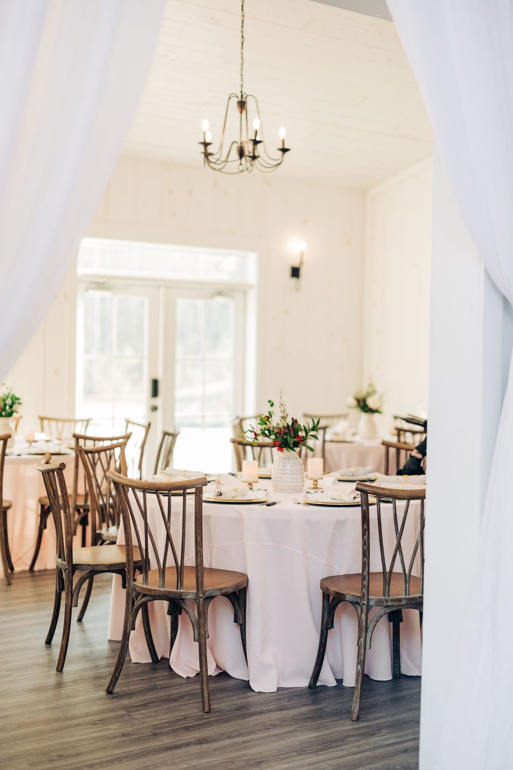 Elegant white reception venue with wooden cross-back chairs and chandeliers in a bright, airy space.
